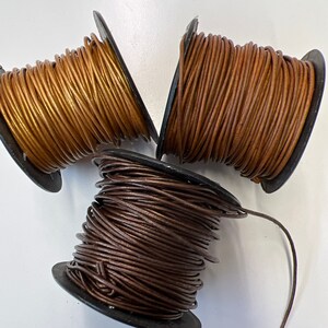 May include: Three spools of leather cord in brown, gold, and dark brown colors. The cord is wound around the spools and is ready to be used for crafting projects.
