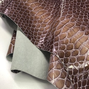 May include: A close-up of a piece of brown leather with a crocodile pattern. The leather is glossy and has a light brown undertone.