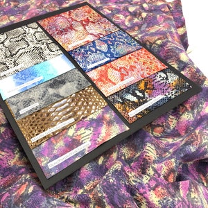 May include: A close-up of a fabric swatch book with different snake skin patterns. The swatches are labeled with the colors: Grey Suede, Metallic Gold/Silver/Bronze, and Ocre/Purple/Pink/Orange.