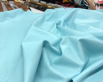 May include: A light blue leather hide, folded and draped on a white surface. The leather is smooth and has a soft, supple texture.