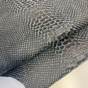 Puede incluir: Primer plano de una tela de piel de serpiente azul oscuro con un patr&oacute;n texturizado. La tela est&aacute; doblada sobre s&iacute; misma, mostrando la textura del material.