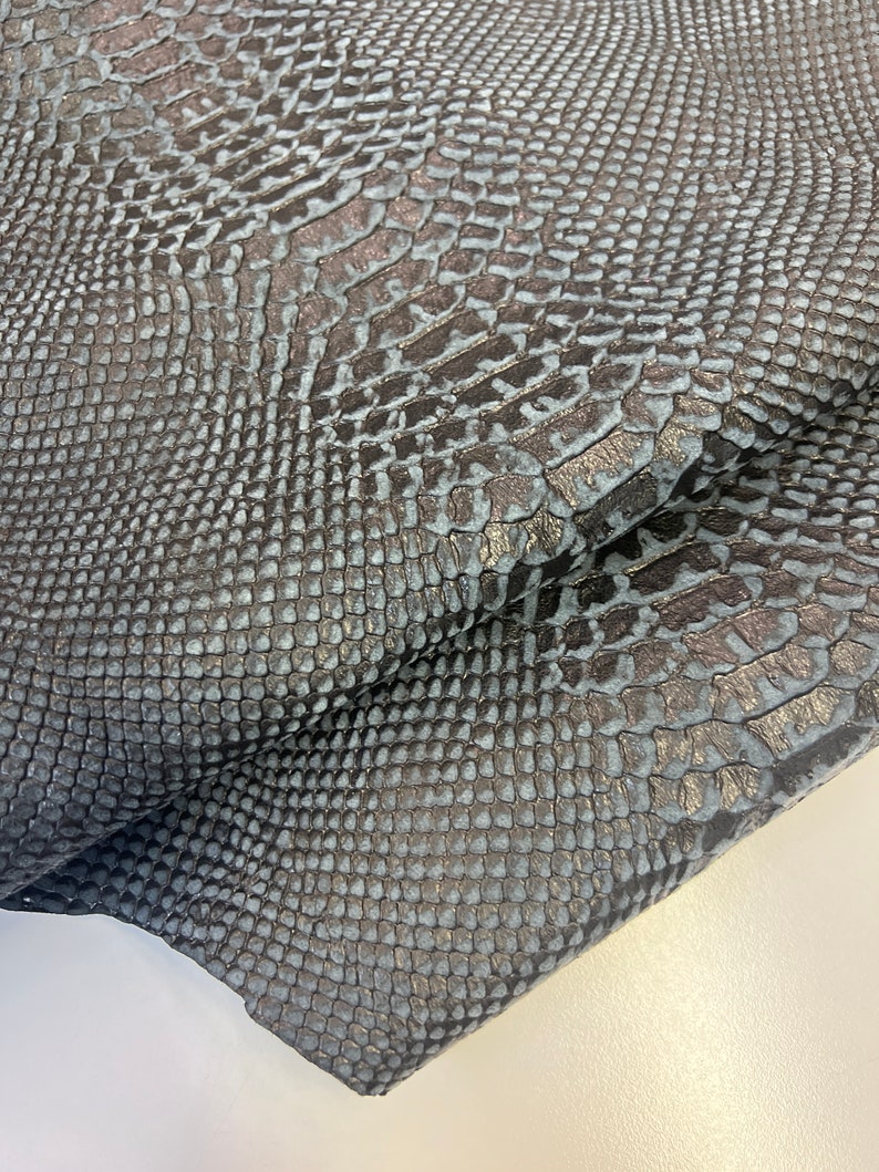 Puede incluir: Primer plano de una tela de piel de serpiente azul oscuro con un patr&oacute;n texturizado. La tela est&aacute; doblada sobre s&iacute; misma, mostrando la textura del material.