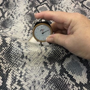 May include: Close-up of a white and black snakeskin fabric with a fabric gauge measuring 0.1mm. The fabric has a textured, scaly pattern.