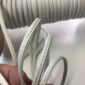 May include: White leather cord with a stitched edge. The cord is rolled up and held in a hand.