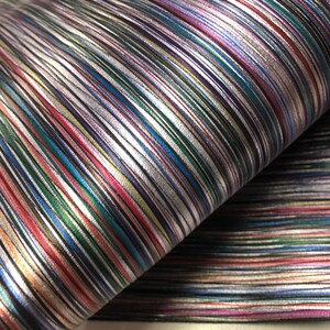 May include: A roll of fabric with a metallic sheen, featuring a pattern of multicolored diagonal stripes. The stripes include silver, red, green, blue, and purple hues. The fabric appears to be folded over itself, revealing its texture and layers.