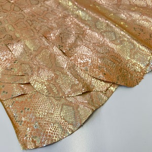 May include: Close-up of a sheet of metallic gold snakeskin leather. The leather has a textured, scale-like pattern and a shiny, iridescent finish. The material is folded over itself, revealing multiple layers.