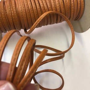 May include: A spool of brown leather cord with a stitched edge. The cord is wound around a cardboard spool.