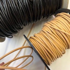 May include: Two spools of leather cord, one black and one beige. The beige cord is unwound and laying on a white surface.