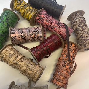 May include: A collection of spools of flat, faux leather ribbon in various colors and snake skin patterns. Colors include green, yellow, black, pink, red, brown, and orange. The ribbon is wound around cardboard spools.