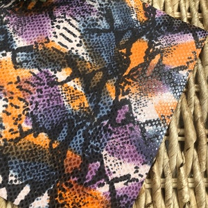 May include: A close-up of a fabric with a snakeskin pattern in shades of orange, purple, black, and white. The fabric has a textured surface.