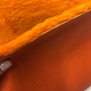May include: Close-up of a piece of orange faux fur fabric with a brown leather backing. The fur is soft and fluffy, and the leather is smooth and textured.