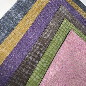 May include: An assortment of textured faux leather sheets in various colors, including gray, gold, purple, brown, green, and pink. The sheets have a reptile-like scale pattern, arranged in a fan shape against a white background.