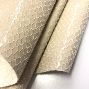 May include: Close-up of a beige snakeskin leather fabric with a textured, embossed pattern. The fabric is folded in thirds, revealing the underside of the material.