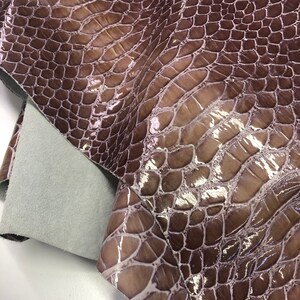 May include: Close-up of a piece of brown leather with a crocodile skin pattern. The leather has a glossy finish.