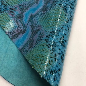 May include: A piece of blue and teal faux snakeskin leather with a glossy finish. The material has a scale pattern and a soft, teal-colored underside. The leather is folded, revealing the texture and color variations.