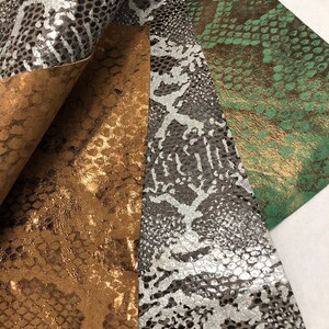 May include: Close-up of several sheets of faux leather with a snakeskin pattern. The colors include metallic gold, silver, black, and green. The sheets are layered, revealing the texture and sheen of the material. Ideal for crafting.