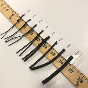 May include: A wooden ruler with black elastic bands of different sizes, measured in millimeters, from 0.5 mm to 6.0 mm.
