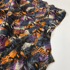 May include: A piece of fabric with a snakeskin pattern in shades of orange, purple, blue, and white. The fabric has a textured surface.