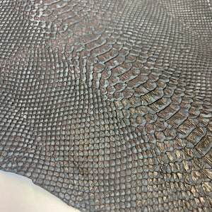 Puede incluir: Un trozo de cuero de serpiente marr&oacute;n oscuro con un brillo sutil. El cuero tiene una superficie texturizada que se asemeja a escamas de serpiente.