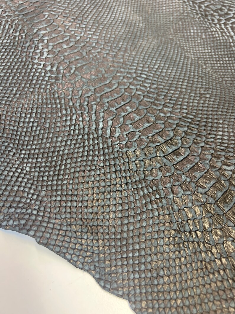 Puede incluir: Un trozo de cuero de serpiente marr&oacute;n oscuro con un brillo sutil. El cuero tiene una superficie texturizada que se asemeja a escamas de serpiente.