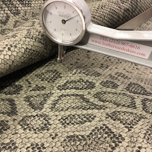 May include: Close-up of a gray snakeskin leather with a gauge measuring the thickness of the material. The gauge reads 0.1 mm and the text on the gauge reads "CALATI MADE IN ITALY". The text "TEL (212) 967-6616 www.leathersuedeskins.com" is also visible.