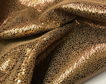 May include: A close-up of a gold leather fabric with a textured, cracked pattern. The fabric has a metallic sheen and is folded in a way that shows the texture.
