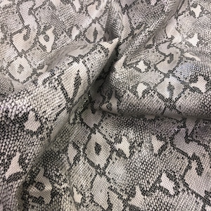 May include: Close-up of a textured fabric with a snakeskin pattern. The material is primarily white, gray, and black, with a repeating geometric design. The fabric is folded, showing depth and texture.