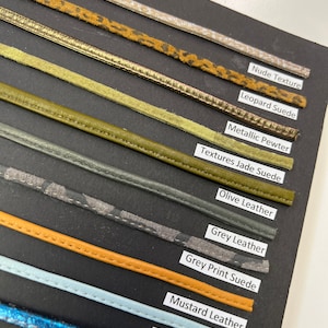 May include: A close-up of a black card with different colored leather trim samples. The trim samples are labeled with names like "Nude Texture", "Leopard Suede", "Metallic Pewter", "Textures Jade Suede", "Olive Leather", "Grey Leather", "Grey Print Suede", "Mustard Leather", and "Baby Blue Leather".