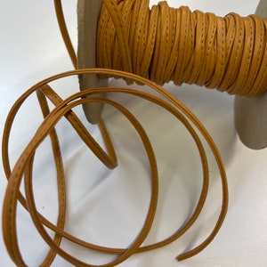 May include: A spool of tan leather cord with a stitched edge. The cord is wound around a cardboard spool.