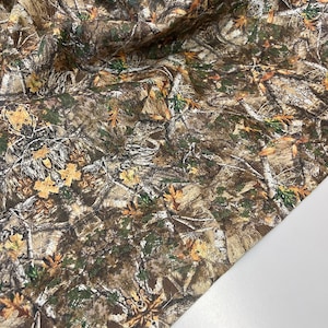 May include: Camouflage fabric with a brown, green, and white pattern. The fabric features a realistic depiction of tree branches, leaves, and bark.