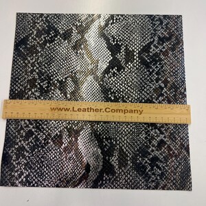 May include: A close-up of a black and silver snakeskin patterned fabric. The fabric has a subtle sheen and a textured surface. The fabric is measured with a ruler that reads "www.Leather.Company".