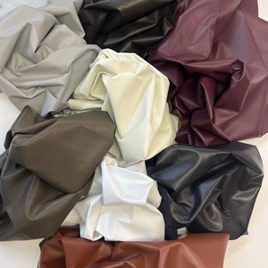 May include: A collection of various colored faux leather fabric swatches. Colors include gray, black, white, brown, and burgundy. The fabric is gathered and folded, showing the texture and sheen of the material. Ideal for crafting or upholstery projects.
