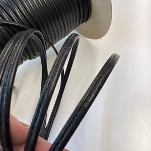 May include: A spool of black leather cord with a stitched edge. The cord is wound around a cardboard spool.