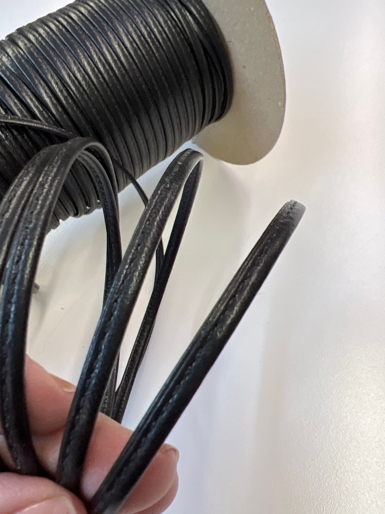 May include: A spool of black leather cord with a stitched edge. The cord is wound around a cardboard spool.