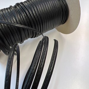 May include: A spool of black leather cord with a textured surface. The cord is wound around a cardboard spool.