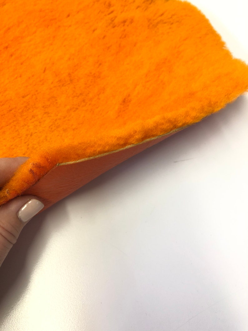 May include: A close-up of a bright orange faux fur fabric with a soft, fuzzy texture. The fabric is folded in half, revealing a smooth, orange backing.