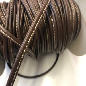 May include: A spool of brown leather cord with white stitching. The cord is rolled up and the end is hanging down.
