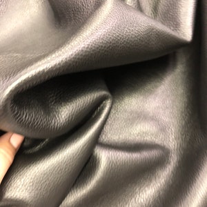 May include: Close-up of a dark gray textured leather fabric. The fabric is folded and has a smooth, shiny surface.