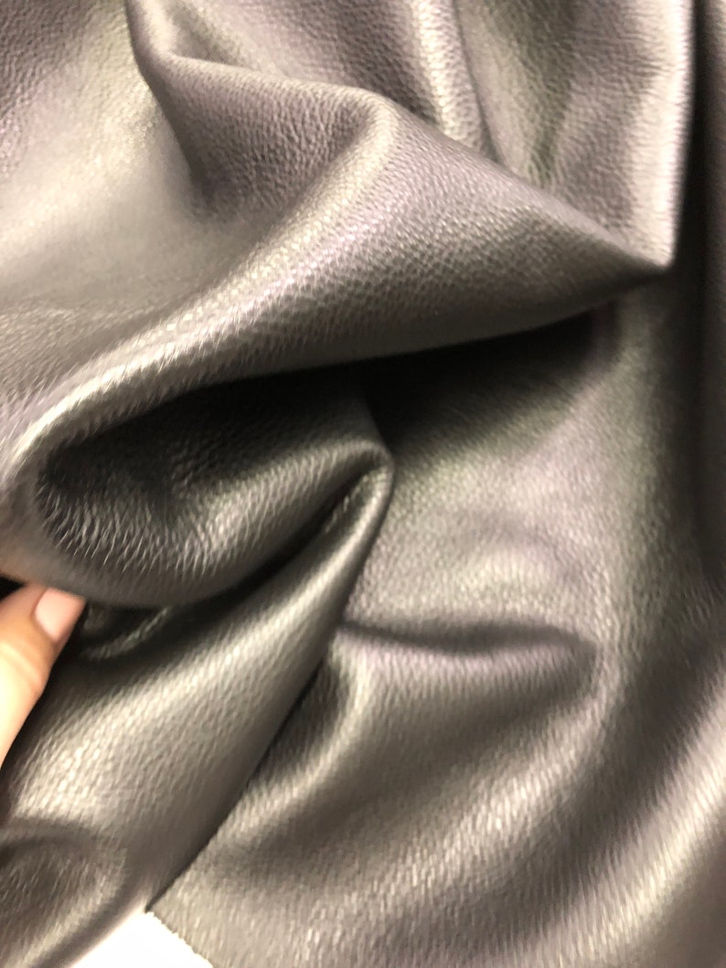 May include: Close-up of a dark gray textured leather fabric. The fabric is folded and has a smooth, shiny surface.