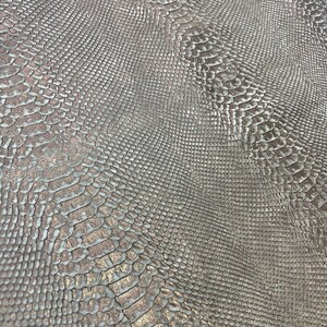 Puede incluir: Primer plano de una tela plateada con relieve de piel de serpiente y un brillo sutil. La tela tiene una superficie texturizada que se asemeja a las escamas de una serpiente.