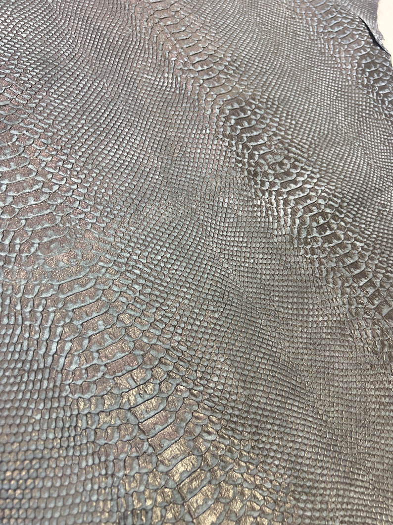 Puede incluir: Primer plano de una tela plateada con relieve de piel de serpiente y un brillo sutil. La tela tiene una superficie texturizada que se asemeja a las escamas de una serpiente.