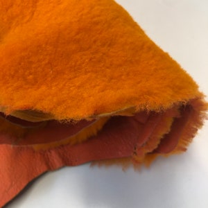 May include: A close-up of a bright orange fleece fabric with a soft, fuzzy texture. The fabric is folded over, revealing a lighter orange underside.