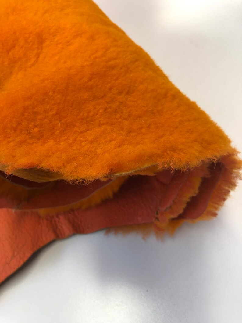 May include: A close-up of a bright orange fleece fabric with a soft, fuzzy texture. The fabric is folded over, revealing a lighter orange underside.