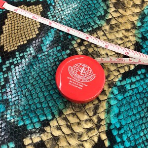 May include: Red tape measure with a white measuring tape and a red lidded container with the text "Leather Suede Skins, Inc." and contact information. The tape measure is on top of a turquoise and black snakeskin pattern.