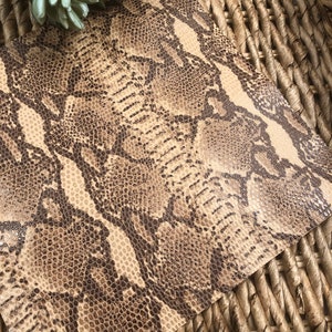 May include: A close-up of a brown and beige snakeskin patterned fabric. The fabric has a textured surface and a subtle sheen.