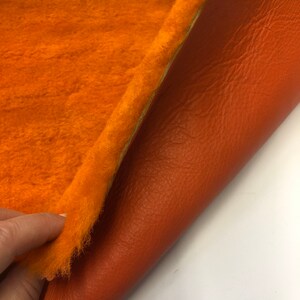 May include: A close-up of a piece of orange faux fur fabric with a brown leather backing. The fabric is soft and fluffy, and the leather is smooth and supple.