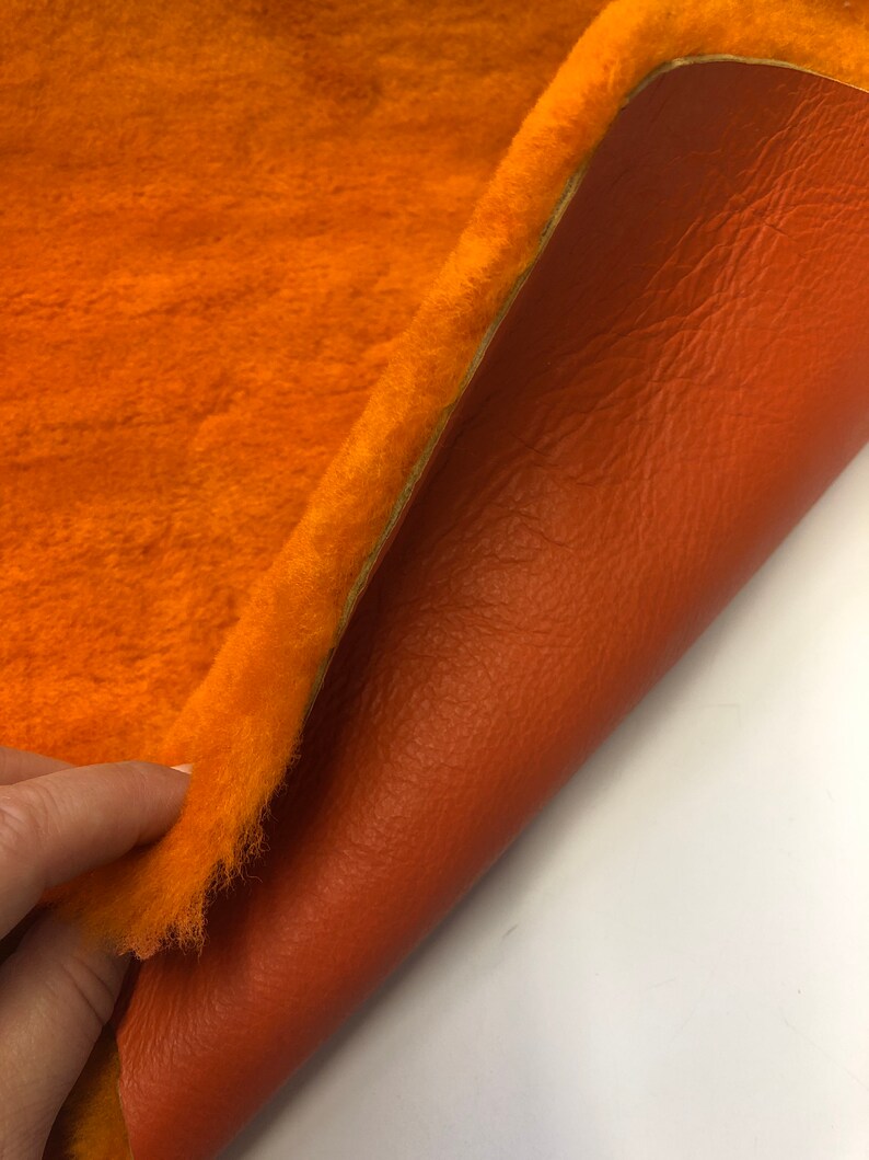 May include: A close-up of a piece of orange faux fur fabric with a brown leather backing. The fabric is soft and fluffy, and the leather is smooth and supple.
