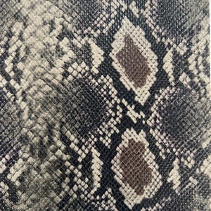May include: A rectangular piece of faux snakeskin fabric. The pattern features a mix of black, gray, white, and brown scales, creating a realistic reptile skin texture. The material appears to be smooth and shiny, suitable for various craft projects.