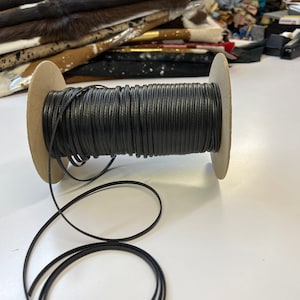 May include: A spool of black leather cord, wound around a cardboard spool. The cord is about 3 mm wide and is ready to be used for crafting projects.