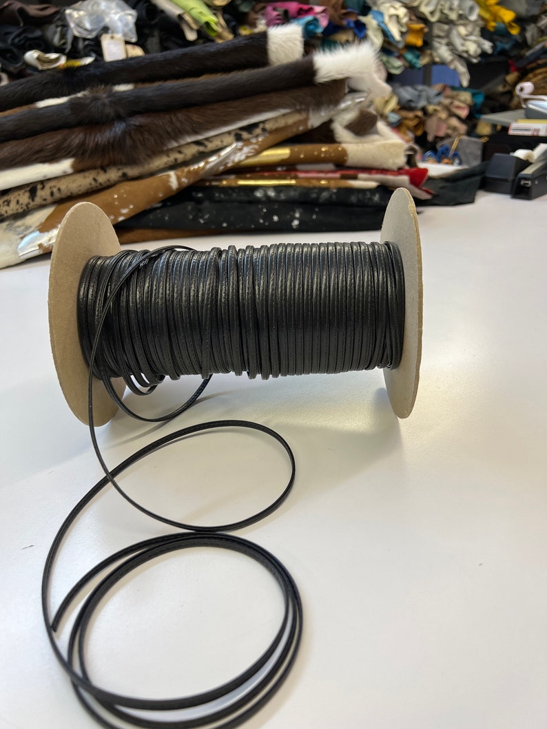May include: A spool of black leather cord, wound around a cardboard spool. The cord is about 3 mm wide and is ready to be used for crafting projects.